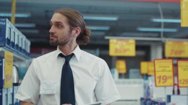 Çekici bir adam, satıcı işi tablet bilgisayar kullanıyor. Süpermarkette teknoloji bölümünde yürüyor. İş dünyasının el alışverişi elektroniği teknolojisi, internet alışveriş merkezi yavaş çekimde.