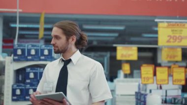 Satıcı yakışıklı süpermarkette teknoloji bölümünde yürüyen tablet bilgisayar kullanıyor. İş dünyasının elektronik el alışverişi, internet teknolojisi, yavaş çekim, yavaş çekimde portre.