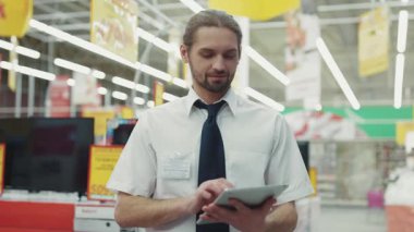 Gülümseyen adam, satış elemanı tablet bilgisayar kullanıyor. Süpermarketin teknoloji bölümündeki kameraya bakıyor. İş dünyasının el alışverişi elektroniği teknolojisi, internet alışveriş merkezinin yavaş çekim portresi.