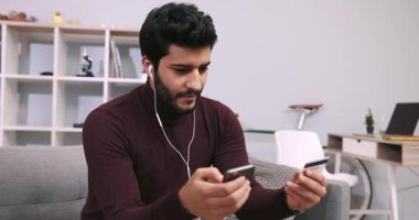 Kulaklıklı çekici genç adam kredi kartını ve telefonunu online alışveriş için kullanıyor, kart numarasını çeviriyor. Aydınlık oturma odası. Para harcamak, kanepe patatesi, tembel alışveriş. Erkek portresi