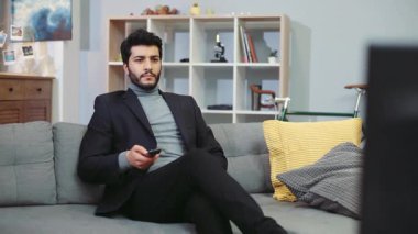 NEW YORK - 5 Nisan 2017: Resmi takım elbiseli çekici genç sakallı adam kanepede oturur, televizyon izler, saati kontrol eder, aceleyle kaçar. Toplantı, başarılı yaşam tarzı, iş. Erkek portresi, kamera sabitleyici.