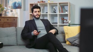 NEW YORK - 5 Nisan 2017: Seksi sakallı adam kanepeye oturur, uzaktan kumandayı kullanır, kanalları değiştirir. Rahatlamış bir atmosfer günü. Dinleniyorum. Yavaş çekim, kamera dengeleyici çekim, erkek portresi.