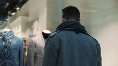 Portre çekici bir adam alışveriş merkezinin Noel yaşam tarzı binasında erkek kıyafetleriyle vitrinlere bakıyor. Noel ağacı mağazasının tasarım parası duruyor. Alışveriş mağazasının vitrininde yavaş çekim.
