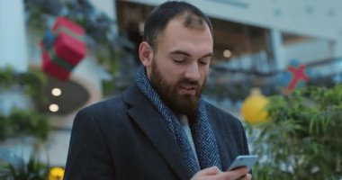 Portre genç çekici adam mutlu olmak için telefon başında duruyor. Yakından alışveriş yapıyor, yeni yıl ışıklarına bakıyor. Yavaş çekimde internet.