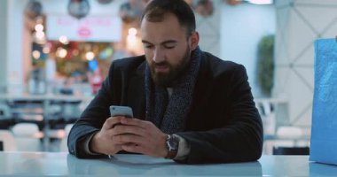 Mutlu yakışıklı genç adam gülümsüyor alışveriş merkezinde oturuyor yakın portre çekici kafe alışverişi bekliyor yeni yıl Masaj ışıkları akıllı telefon mobil internet yavaş çekim