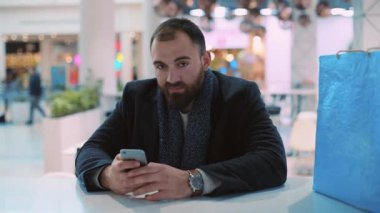 Genç adam telefonuyla oturuyor kameraya bakıyor gülümsüyor omuz silkiyor, mutlu oluyor, yakından bakıyor, alışveriş yapıyor, alışveriş yapıyor, yeni yıl resmine bakıyor, akıllı telefon interneti yavaş çekim yapıyor.