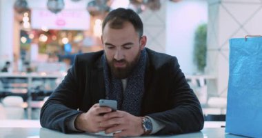 Kapalı bir yüz genç çekici adam oturuyor kameraya bakıyor gülümsüyor mutlu portre yakışıklı alışveriş bekliyor yılbaşı akıllı telefon ışıkları internet yavaş çekim