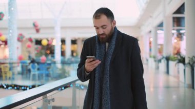 Çantalı genç ve çekici bir adam alışveriş merkezinde gülümsüyor mutlu portre yakışıklı alışveriş bekliyor yılbaşı cep telefonu akıllı telefon günlük internet yavaş çekim