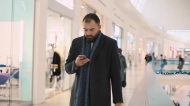Gülümseyen genç ve çekici bir adam telefon kullanıyor. Alışveriş merkezinde duruyor. Mutlu portre yakışıklı, alışveriş bekliyor.