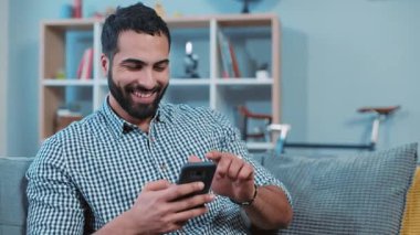 Mutlu genç adam telefonda mesaj yazıyor, gülümsüyor. Kanepeye oturup ev portresi çekiyor. Akıllı telefon ellerini kapatıyor, elektronik uygulama kullanıyor.