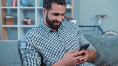 Çekici genç adam telefonu kullan gülümse mutlu hisset kanepeye otur ev portresi kapat akıllı telefon elleri elektronik uygulama mesajı yaz eğlence yaşam tarzı iletişimi dokun