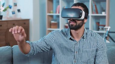 Çekici bir adam sanal gerçeklik gözlüğü kullanıyor koltukta oturuyor ellerini sallıyor ev portresinde gülümsüyor oyun evini kapıyor 3 boyutlu internet teknolojisi yeniliği ağır çekim