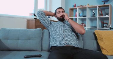 Uykulu, yorgun, yakışıklı, kanepede oturan genç adam elinde uzaktan kumandayla televizyona bakıyor.