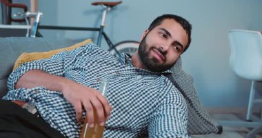 Rahatla genç, gülümseyen, bira içen, bira içen, uzaktan kumandayı evde tek başına bir kanepede tutan, içki içen, mutlu bir hayat tarzı olan, televizyon seyreden, ağır çekimli, mutlu bir portre.