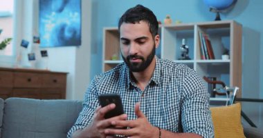 Çekici genç adam, gülümse. Kanepeye otur. Ev portresi, akıllı telefon, elektronik uygulama.