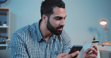 Çevrimiçi satın alan gülümseyen genç adam telefon ve kredi kartı kullanıyor. Kanepe üzerinde alışveriş cep telefonu müşterisi. Kapalı telefon.