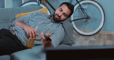 Gülümseyen, bira içen, televizyon seyreden, uzaktan kumandayı evde bir koltukta tutan, portre çeken, genç, yalnız şişeci, kanepede mutlu, alkollü bir hayat süren, ağır çekimde televizyon izleyen çekici bir adam.