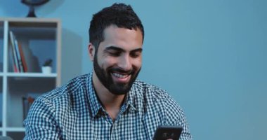 Gülümseyen genç adam telefonu kullan. Mutlu ol. Kanepeye otur. Ev portresi, akıllı telefon, elektronik uygulama.