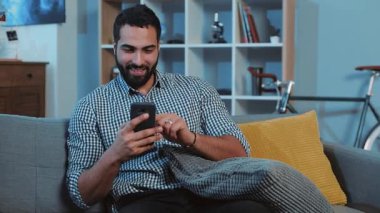 Gülümseyen mutlu genç adamın portresi telefon kullan. Kanepeye otur. Akıllı telefon, elektronik uygulama.