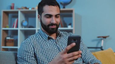 Mutlu ve çekici genç adam gülümsüyor. Kanepeye otur. Akıllı telefon, elektronik uygulama, mesaj yaz.