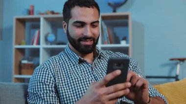 Sevimli, mutlu, çekici genç adam gülümsüyor. Kanepeye otur, akıllı telefon, elektronik uygulama. Mesaj yaz.