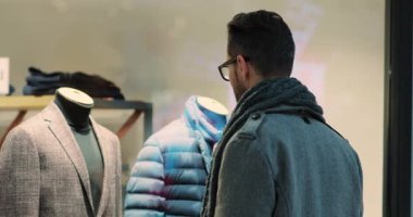 Genç ve çekici bir adam alışveriş merkezinin Noel yaşam tarzı binasında erkek kıyafetleriyle vitrinlere bakıyor. Noel ağacı mağazasının tasarım parası duruyor. Alışveriş mağazasının vitrininde yavaş çekim.