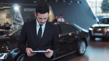 Kravat tutan, elinde belge tutan ve araba galerisindeki arabaların arka planında omuz silken sempatik bir iş adamı..