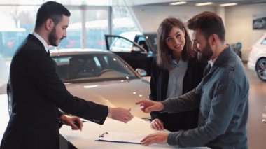 Karı koca araba alıyor. Çekici genç adam araba belgelerine imza atıyor, satıcıyla tokalaşıyor. Mutlu çift kucaklaşması. Araba galerisi.