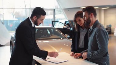 Satıcı kontrat imzalıyor, kalemi adama veriyor. Genç adam belgeye imza atıyor. Satıcı yeni arabaların arka planında arabanın anahtarlarını veriyor..