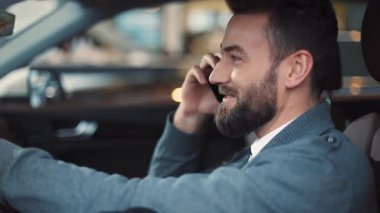 Sakallı, şoför koltuğunda oturan ve cep telefonuyla konuşan yakışıklı bir adam. Mavi kazaklı genç adam gülümsüyor, akıllı telefon kullanıyor..