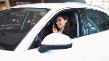 Çekici esmer, yeni beyaz arabanın sürücü koltuğunda oturuyor. Direksiyon başında genç bir kız. Araba galerisi.