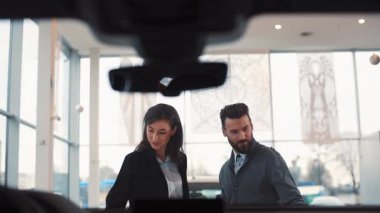 Güzel bir çift araba galerisinde araba seçiyor. Takım elbiseli hoş bir satış danışmanı genç insanlara yaklaşıyor ve araba modelinden bahsediyor. Gülümsüyor.