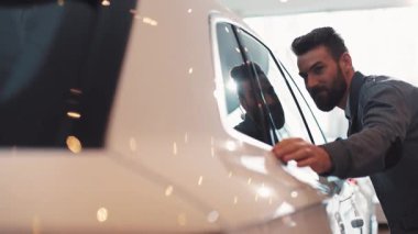 Sakallı genç adam araba galerisinde arabaya dikkatlice bakıyor. Parlak beyaz bir arabanın yan camına dokunan adam..