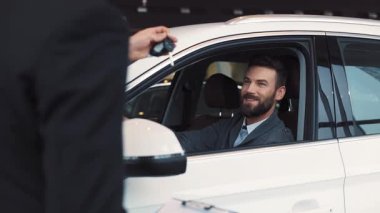 Genç yakışıklı işadamı beyaz arabada sürücü koltuğunda oturuyor. Satıcı araba galerisinin alıcısına anahtarları veriyor. Beyaz adam dikiz aynasına bakıyor..