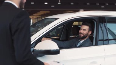 Prestijli araba galerisinde direksiyonda oturan mutlu çekici adam. Siyah takım elbiseli satıcı adama anahtarları verip gidiyor..