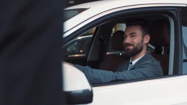 Yeni beyaz arabasında oturan sakallı genç adam. Adam gülüyor ve anahtarlar için satıcı-danışmana teşekkür ediyor. Erkekler konuşuyor, gülüyor..