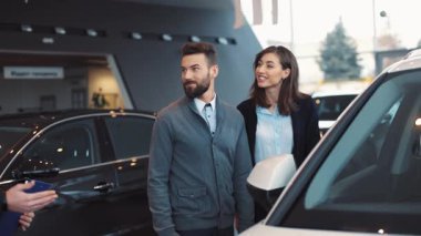 Genç evli çift, prestijli araba galerisinde düğünden sonra araba seçiyor. Siyah takım elbiseli satıcı sevilen modelin detaylı tarifini anlatıyor.