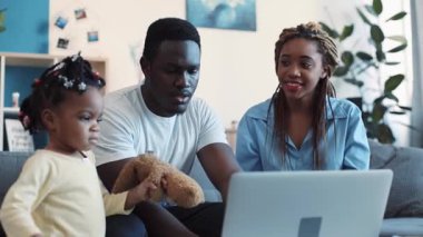 Genç Afrikalı Amerikalı baba dizüstü bilgisayarı koltukta otururken kullanıyor, karısı da onu ve tatlı kızlarını oyuncak ayıyla oynarken izliyor. Aile hayali, ortaklık. Yavaş çekim, kamera dengeleyici çekim
