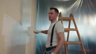 Yakışıklı, sıradan görünümlü genç adam aktif olarak duvarı beyaz bir boyayla boyuyor. Tahta merdiven, arka planda lastik çarşaf. Pozitif ruh hali, neşeli duygular. Yavaş çekim, kamera dengeleyici çekim