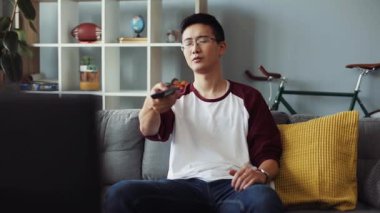 Gerçekten çekici, gözlüklü genç Çinli bir adam kanepede oturur ve TV 'yi açar ev bakımı teknolojisi erkek işi sıkıcı video kanepede ağır çekimde portre yakın çekim