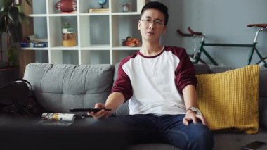 Gözlüklü genç çekici Çinli adam kanepede oturur ve TV 'yi açar. Ev bakımı teknolojisi erkek işi. Boş zamanlardan sıkılan video kanepe portresi.