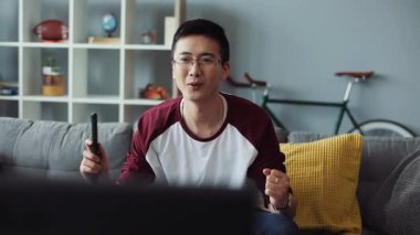 Mutlu gülümseyen çekici gözlüklü genç Çinli adam kanepede oturur ve TV 'yi açar ev bakımı teknolojisi erkek işi sıkıcı video kanepede ağır çekim portresi kapar.