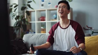 Sevimli, gülümseyen, gözlüklü genç Çinli adam kanepede oturur ve TV 'yi açar. Mutlu görünümlü ev teknolojisi erkek işi. Keyif çatan video kanepe portresi.