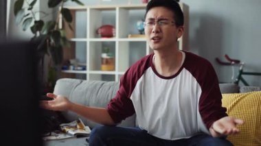 Gergin, çekici, gözlüklü genç Çinli adam kanepede oturur ve TV 'yi açar. Ev arama teknolojisi. Hüzünlü erkek işi.