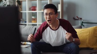 Gülümseyen çekici, gözlüklü genç Çinli adam kanepede oturur ve TV 'yi açar ev arama teknolojisi erkek işi sıkıcı video kanepede ağır çekim portresi.