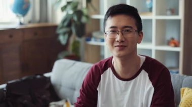 Portre güzel, çekici, çekici, gözlüklü, başarılı Çinli adam kameraya bakıyor, kanepeye oturuyor, gülümsüyor, Asyalı, yakışıklı, yakışıklı iş adamı, yavaş çekimde, neşeli, yakın çekimde.