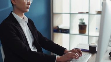 Ciddi genç Çinli gözlüklü adam ofis işindeki ekrana bakmak için bilgisayar kullanıyor. İnternetle yüz yüze, teknoloji işadamı, iş arkadaşı, çekici iç mekan yakışıklı, ağır çekim.