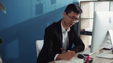 Gözlüklü genç Çinli adam deftere yazıyor. Ofis işindeki ekran bilgisayarına bak. İnternete bak.