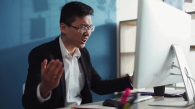 Üzgün, gözlüklü genç Çinli adam ofis işindeki ekrana bakmak için bilgisayar kullanıyor. İnternet yüz teknolojisi. Gergin, dizüstü bilgisayar işadamı.