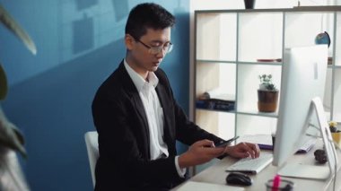 Gözlüklü genç Çinli adam telefonda bilgisayar kullanıyor. Ofiste ekrana bak. İnternet yüz teknolojisi iş adamı.